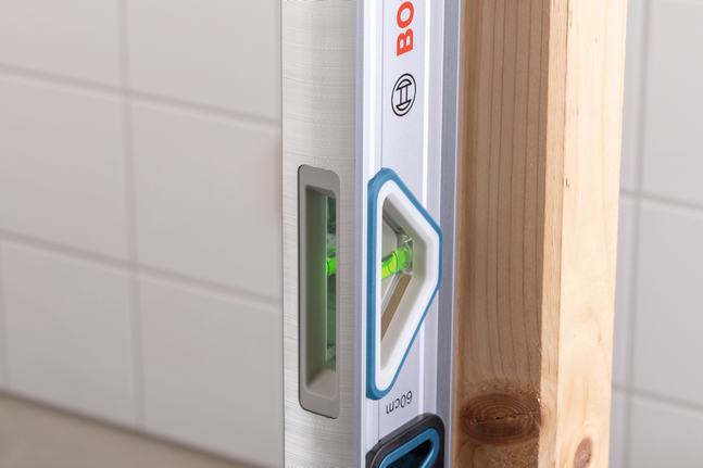 Vodna tehtnica preverja navpično poravnavo lesenega tramca ob steni s ploščicami.